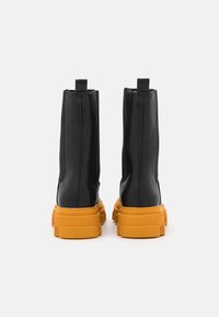 Une paire de bottes en cuir noir mi-mollet avec des semelles en caoutchouc orange vif, épaisses et texturées, vues de l'arrière sur un fond blanc.