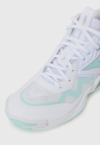 Chaussure de sport blanche en mesh et matériaux synthétiques, avec des accents vert clair, des motifs texturés et un système de laçage traditionnel.
