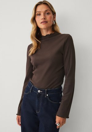 Kvinde med blondt hår iført en brun langærmet top og mørkeblå jeans med høj talje står foran en ensfarvet lys baggrund.