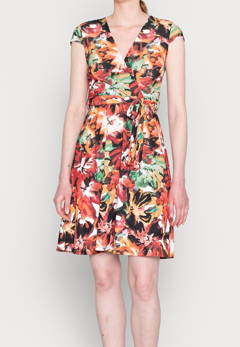 Femme portant une robe sans manches, longueur genou, à motifs floraux aux couleurs rouge, orange, vert et noir, avec une ceinture nouée à la taille.