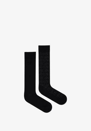 Dwie czarne skarpetki załogi, jedna gładka, a druga we wzór z powtarzającym się tekstem "ARMANI" w ciemnoszarym kolorze, pokazane obok siebie na białym tle.