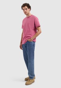 Rosa Baumwoll-T-Shirt mit Rundhalsausschnitt und kurzen Ärmeln, kombiniert mit lockeren blauen Jeans und beigen Schnürschuhen. Einfache Logo-Details sind sichtbar.