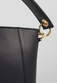 Sort lædertaske med et elegant design, der har en guldfarvet metalring og stikningsdetaljer på siden. Justerbar skulderrem medfølger.
