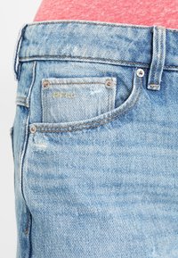 Vaaleansinisiä farkkuja, joissa on kulutettu pinta. Takapocketissa on keltaista ommelta ja metallinen niitti. Kankaan pinta on tekstuuri ja hieman kulunut.