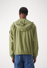 Giacca a vento verde oliva chiaro con cappuccio, cordoncino, polsini ed elastico in vita; tessuto liscio, indossata sopra pantaloni corti in denim blu.