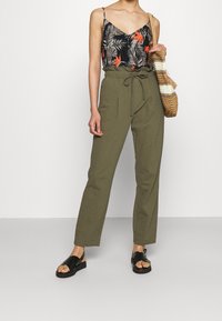 Top negro con estampado floral de hojas rojas y verdes, pantalones de cuerda color verde oliva y sandalias negras con una tira texturizada. Bolso tejido en la mano.