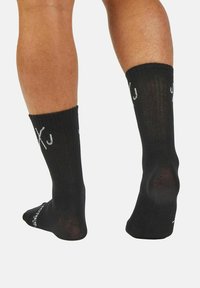 Black socks made of a textured fabric, featuring a ribbed cuff and white logo detailing on the sides, showing heel and ankle area.