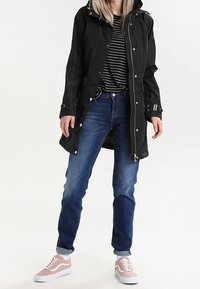 Cappotto nero impermeabile con cappuccio, hardware color argento e chiusura a scatto. Abbinato a jeans blu e sneakers rosa con dettagli bianchi.