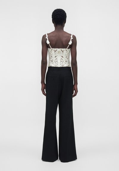Γυναίκα που στέκεται πλάτη, φορώντας λευκό μανίκωμα μπλούζας με δαντέλα και λουλουδάτες λεπτομέρειες και μαύρο φαρδύ παντελόνι μπροστά σε απλό φόντο.