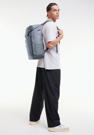 Mochila gris con un diseño estructurado, que cuenta con un zipper frontal y correas ajustables. Modelo que lleva una camisa gris y pantalones negros.