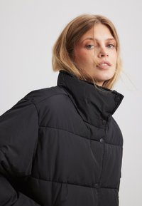 Melnā pildītā jaka ar augstu apkakli, spiedpogu aizdari un izšūtu tekstūru. Audums izskatās gluds un viegls.