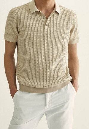 Homme portant un polo beige à manches courtes en maille torsadée avec un pantalon blanc, les mains posées de manière décontractée dans les poches, debout devant un fond uni.