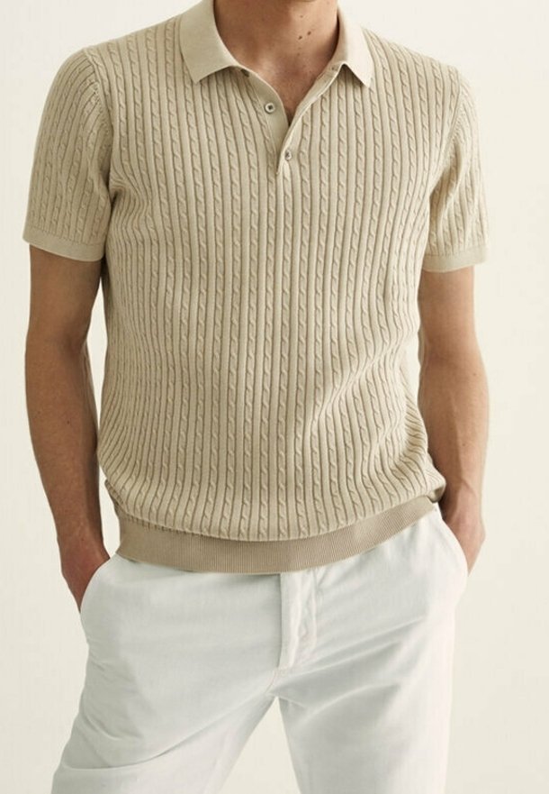 Homme portant un polo beige à manches courtes en maille torsadée avec un pantalon blanc, les mains posées de manière décontractée dans les poches, debout devant un fond uni.