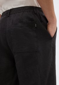 Personne portant un pantalon noir ample avec poches arrière et latérales, main dans la poche latérale, chemise blanche rentrée, étiquette Ecoalf visible sur la poche.