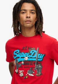 Rød bomulds T-shirt med tegneserie grafikdesign og "SuperDry" tekst, der viser karakterer og urbane elementer. Korte ærmer, rund halsudskæring.