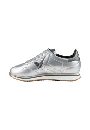Zapatilla deportiva plateada de caña baja con cordones grises, forro interior negro y suela blanca con dibujo marrón.