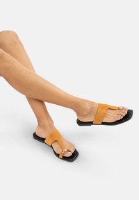 Oranje suède sandalen met een zwarte platte zool, voorzien van een T-vormige band, metalen accent en open teen ontwerp.
