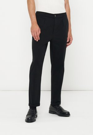 Homme portant un pantalon noir slim avec des ourlets revers et des mocassins en cuir noir, debout devant un fond blanc.
