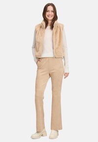 Kamizelka z sztucznego futra w kolorze beżowym z zamkiem z przodu, zestawiona z dopasowanymi spodniami przypominającymi zamsz w kolorze beżowym. Model nosi biały sweter i jasne botki.
