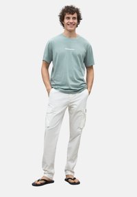 Camiseta de algodón verde claro con texto blanco, combinada con pantalones cargo blancos con bolsillos laterales, y chanclas negras.