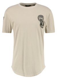 Beige cotton t-shirt with short sleeves, rounded hem, and two black patches featuring text and emblem details. Standard crew neck design.
