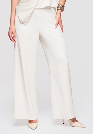 Kvinde iført hvide bukser med vide ben, beige spidse høje hæle og gyldne armbånd på højre håndled mod en ensfarvet baggrund.