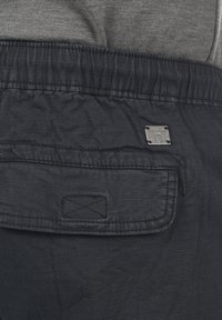 Shorts dintr-un material închis, cu o suprafață texturată, talie elastică și un buzunar posterior cu un accent metalic în formă de logo.