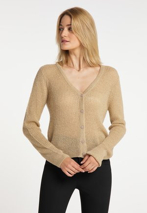 Woman with shoulder-length blonde hair wearing a beige buttoned knit cardigan and black pants standing against a plain white background.