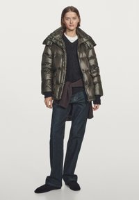 Młoda kobieta w błyszczącej oliwkowej kurtce puchowej, czarnym swetrze, niebieskich dżinsach i czarnych butach, stojąca na jednolitym tle.