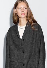 Szary, pleciony kardigan z kołnierzem szalowym, z trzema czarnymi guzikami, dwoma bocznymi kieszeniami i luźnym krojem. Widoczne detale strukturalne materiału.
