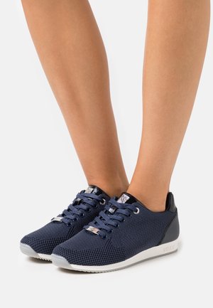 Zapatillas de punto azul marino con detalles negros en el talón, suelas grises y cordones, usadas en pies descalzos sobre un fondo blanco.