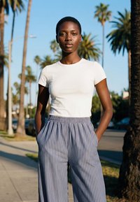 Young woman with short hair wearing a white t-shirt and blue striped pants stands with hands in pockets on a palm tree-lined street.