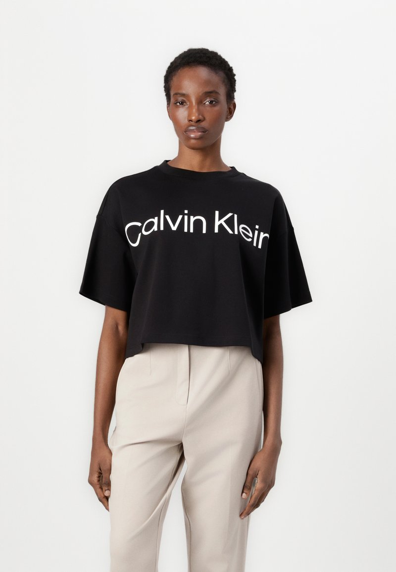 Black cropped t-shirt with “Calvin Klein” logo in white. Features short sleeves and a relaxed fit, paired with light-colored pants.