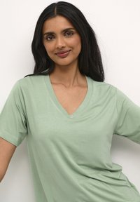 Tricou scurt cu mânecă scurtă, de culoare verde deschis, cu decolteu în V, fabricat dintr-un material moale; are o croială lejeră și o textură netedă, cu cusături minime.