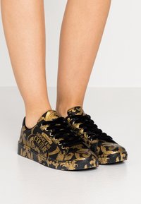 Černé tenisky se zlatým květinovým vzorem, kulatou špičkou, plochou podrážkou a černými tkaničkami. Obsahují značku "Versace Jeans Couture".