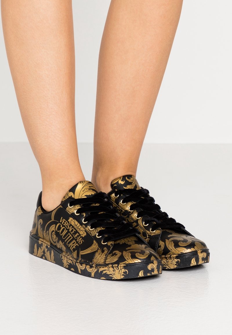 Černé tenisky se zlatým květinovým vzorem, kulatou špičkou, plochou podrážkou a  černými tkaničkami. Obsahují značku "Versace Jeans Couture".