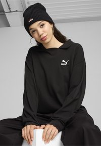 Black oversized long-sleeve polo with a collar, featuring a white Puma logo. Worn with a black ribbed beanie and relaxed black pants.