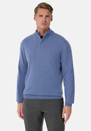 Helles blauer Pullover mit halbem Reißverschluss, gerippten Bündchen und Saum, aus weichem Stoff. Slim Fit, getragen über einem dunklen Unterhemd.