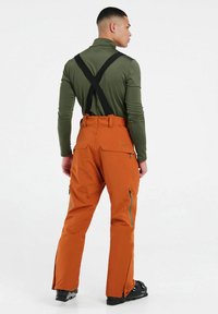 Heren sneeuwbroeken in oranje, gemaakt van waterdicht materiaal, hebben verstelbare zwarte bretels, ritsen op de benen en een loose fit voor bewegingsvrijheid.