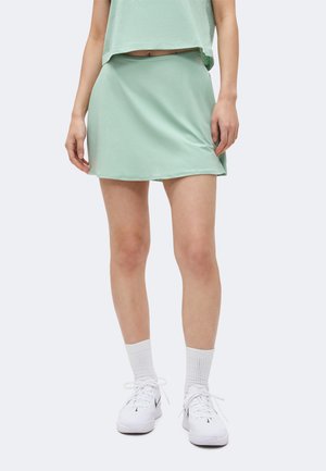 Person iført et lysegrønt nederdel, matchende crop top, hvide crew sokker og hvide Nike-sneakers, stående på en ensfarvet baggrund.