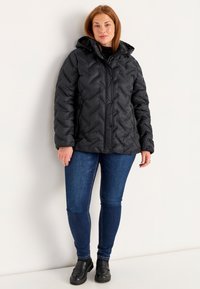 Svart pufferjacka med ett chevronmönster, dragkedjestängning, knappdetaljer och huva. Parat med blå jeans och svarta slip-on skor.
