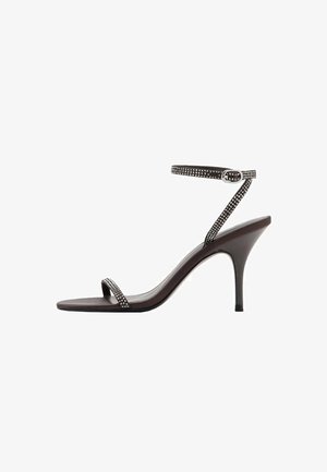 Černé sandály na vysokém podpatku s hladkým povrchem, tenkým popruhem kolem kotníku a stříbrnou sponou. Obsahují úzký popruh přes prsty, ozdobený cvočky.