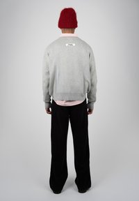 Personne portant un bonnet rouge en tricot, un pull gris avec une étiquette dans le dos, une chemise rose à rayures et un pantalon noir à jambes larges, debout de dos.