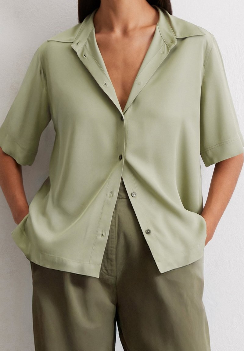 Chemise à manches courtes, boutonnée, de couleur vert clair, en tissu lisse. Elle possède un col et des poches, portée avec un pantalon ample de couleur olive.