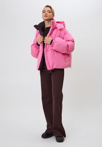 Žena v ružovej prešívanej bunde cez čierne tričko, hnedé zvonové nohavice a čierne topánky stojí na bielom pozadí.