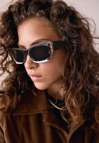 Schwarze und schildpattfarbene Sonnenbrillen mit Cat-Eye-Form, auffälligen Fassungen und dunklen Gläsern, kombiniert mit einer braunen Oberbekleidung und strukturiertem Haar.