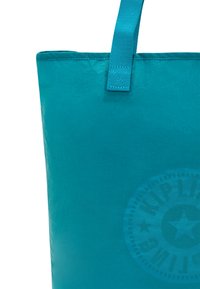 Blå tote-väska med en glänsande yta, som har en robust tygdesign, en förstärkt rem och en subtil cirkulär logodetalj.