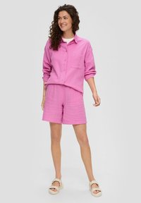 Roze overhemd met knoopjes en een zak, en bijpassende shorts, gemaakt van een gestructureerde stof. Gepaard met lichtgekleurde plateausandalen.