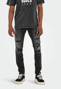 Jean denim noir avec des détails déchirés, associés à un t-shirt graphique gris et des baskets noires avec des semelles blanches.