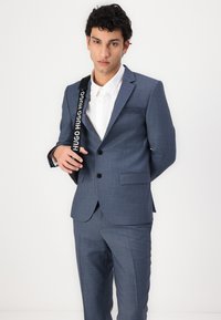 Blauer strukturierter Anzug mit taillierter Silhouette, einknopfiger Verschluss und Reverskragen. Kombiniert mit einem weißen Hemd und einem schwarzen Markenriemen.